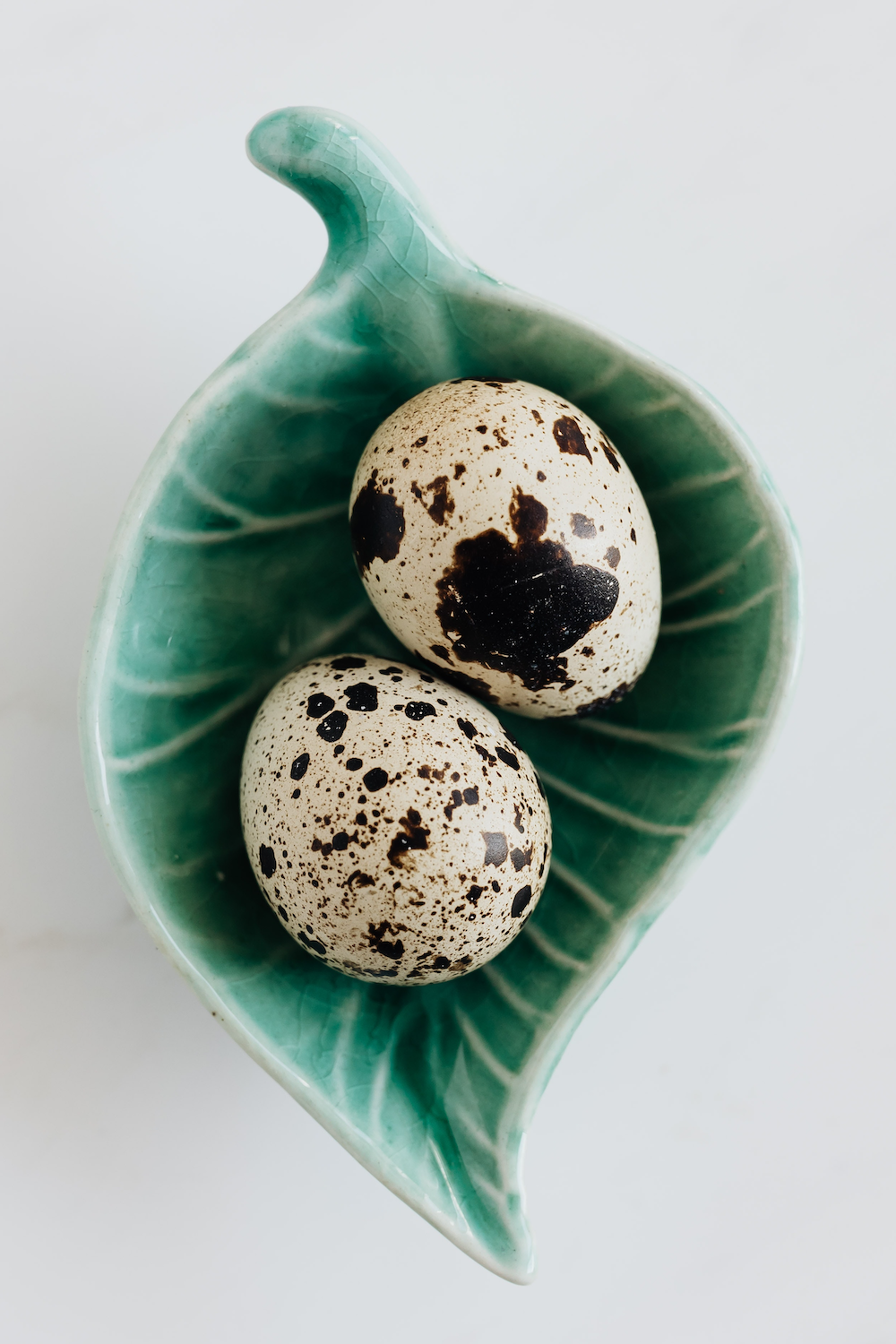 Two white-with-black-spotted quail eggs in a ceramic dish shaped like a green leaf.