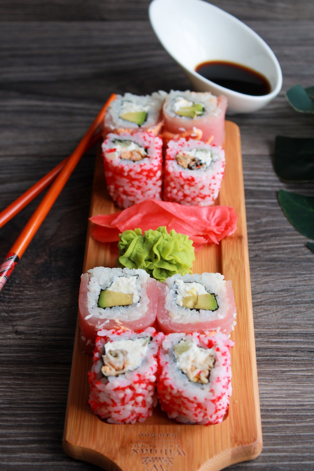wasabi paste and ginger slices surrounded by sushi on a wooden platter, next to a pair of red chopsticks and a small bowl of soy sauce