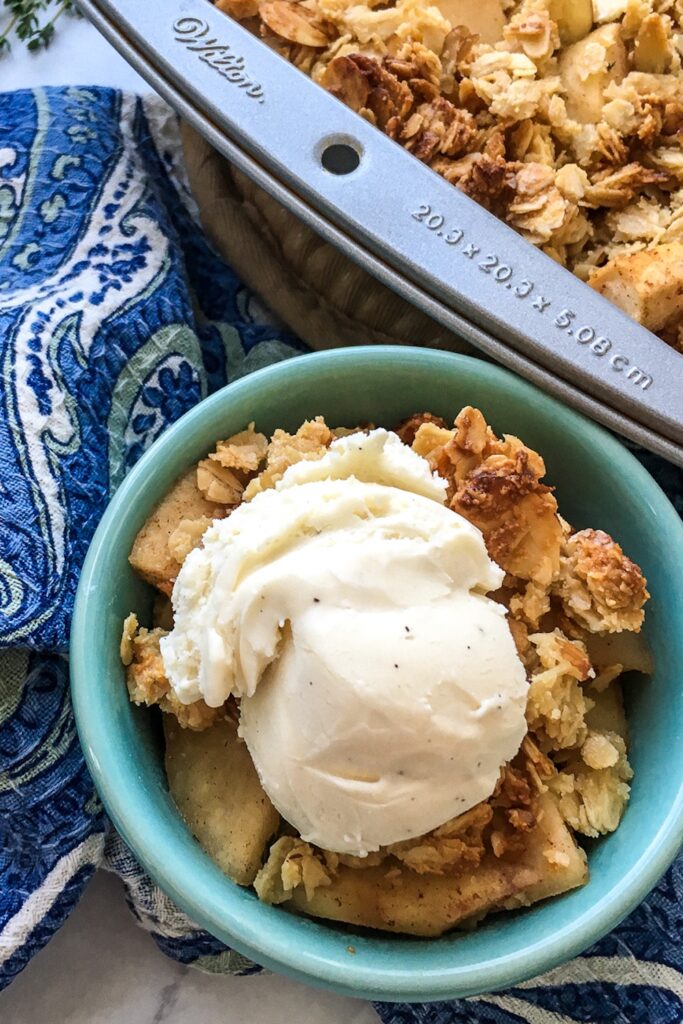gluten free apple crisp in a bowl topped with ice cream, sitting next to a pan of apple crisp