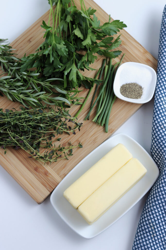 rosemary, chives and parsley on a cutting board next to two sticks of butter on a plate
