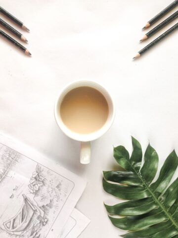 top view of cup of coffee surrounded by pencils, a drawing and a green tropical leaf