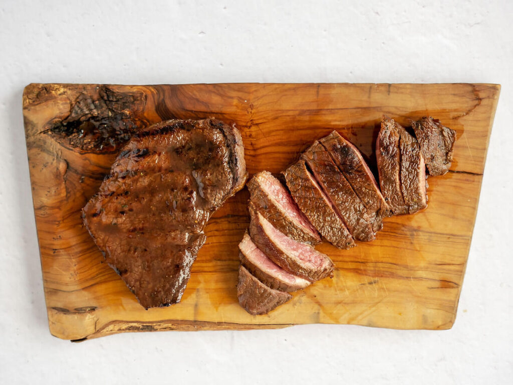 Top view of a cooked steak and a sliced cooked balsamic marinated steak on a wooden cutting board.