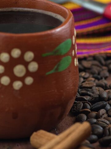 Mexican coffee in a jarrito next to piloncillo and cinnamon sticks