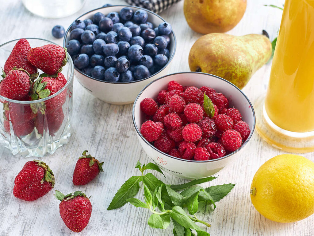 assorted berries in bowls surrounded by scattered berries, pears, lemon and mint leaves