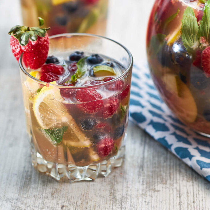 fruit filled glass of mint berry sangria garnished with a strawberry next to a glass pitcher of mint berry sangria