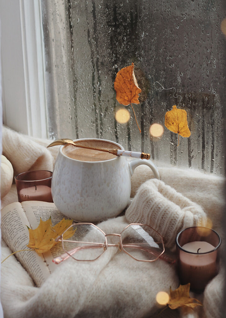 filled earthenware ceramic mug with coffee and a spoon balanced on top, in fornt of a window and scattered with fall leaves