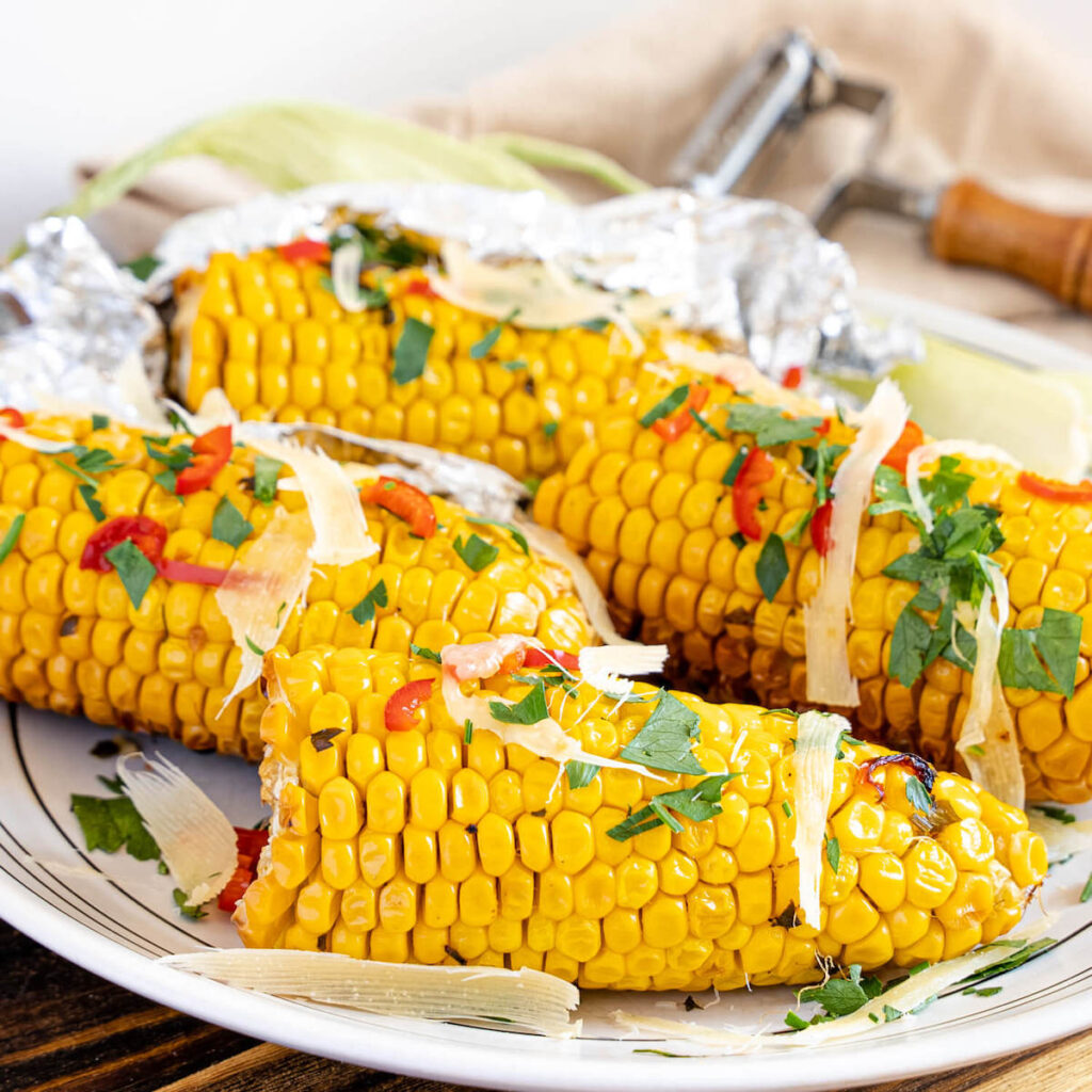 grilled corn on the cob on a plate and garnished with red chili slices, chives, and shaved parmesan cheese