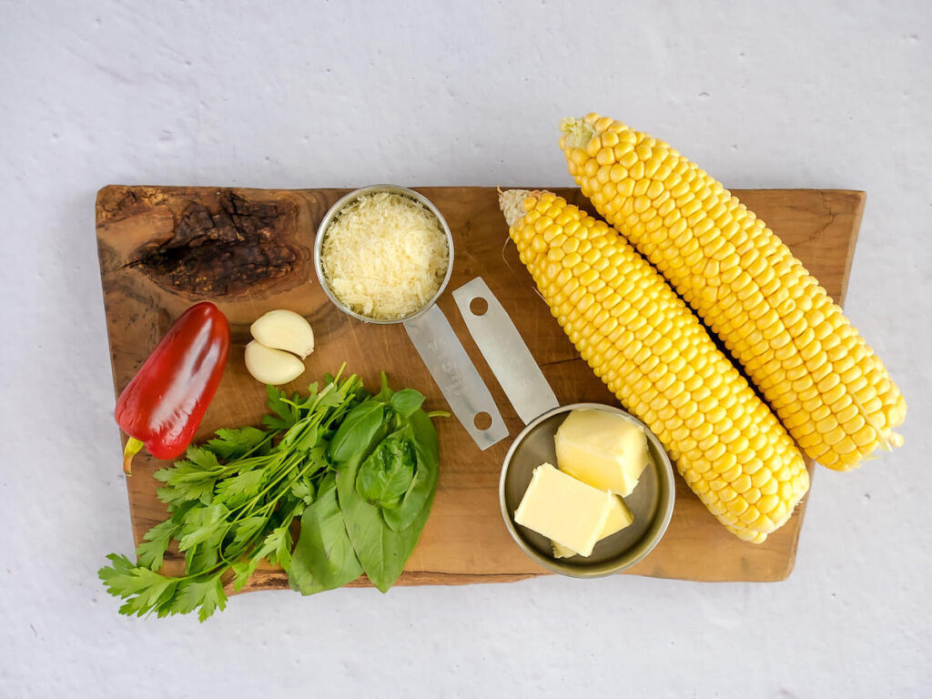 two corn on the cob, fresh basil, parsley and red chili, a cup with butter and a cup with grated parmesan cheese displayed on a wooden cutting board
