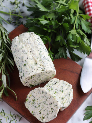 sliced compound herb butter log on a cutting board surrounded by fresh green herbs