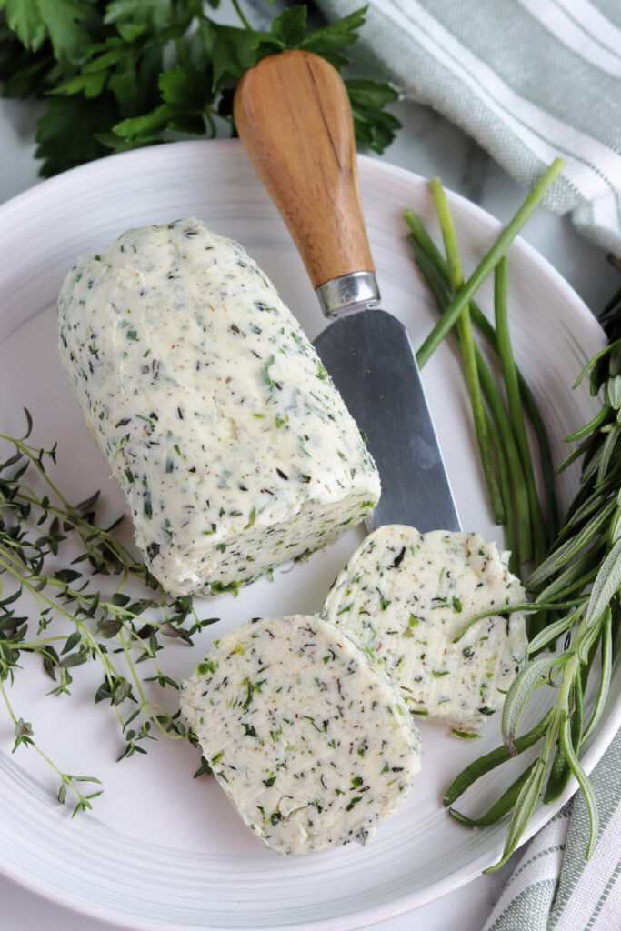 compound herb butter log with a few slices on a white plate with a butter knife and fresh thyme, rosemary and chives