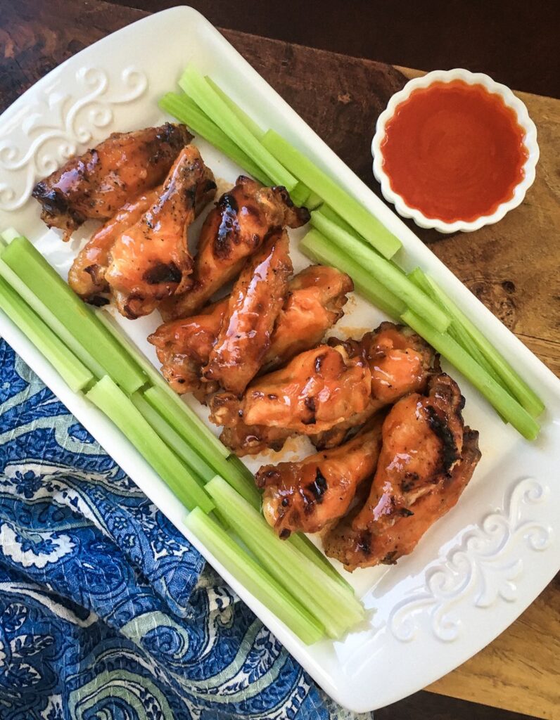 buffalo chicken wings surrounded by cut celery on a white plate next wo a small bowl of sauce