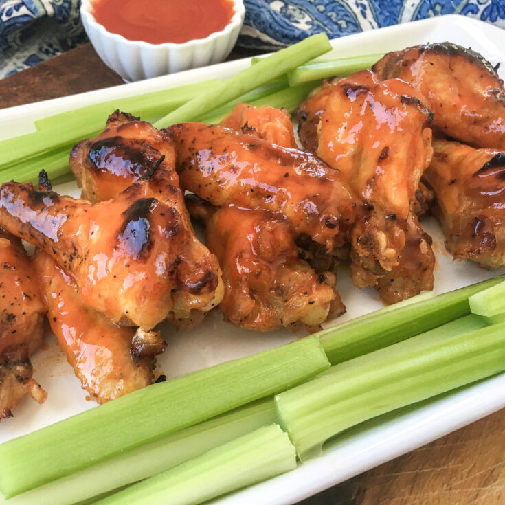 buffalo chicken wings surrounded by cut celery on a white plate next wo a small bowl of sauce
