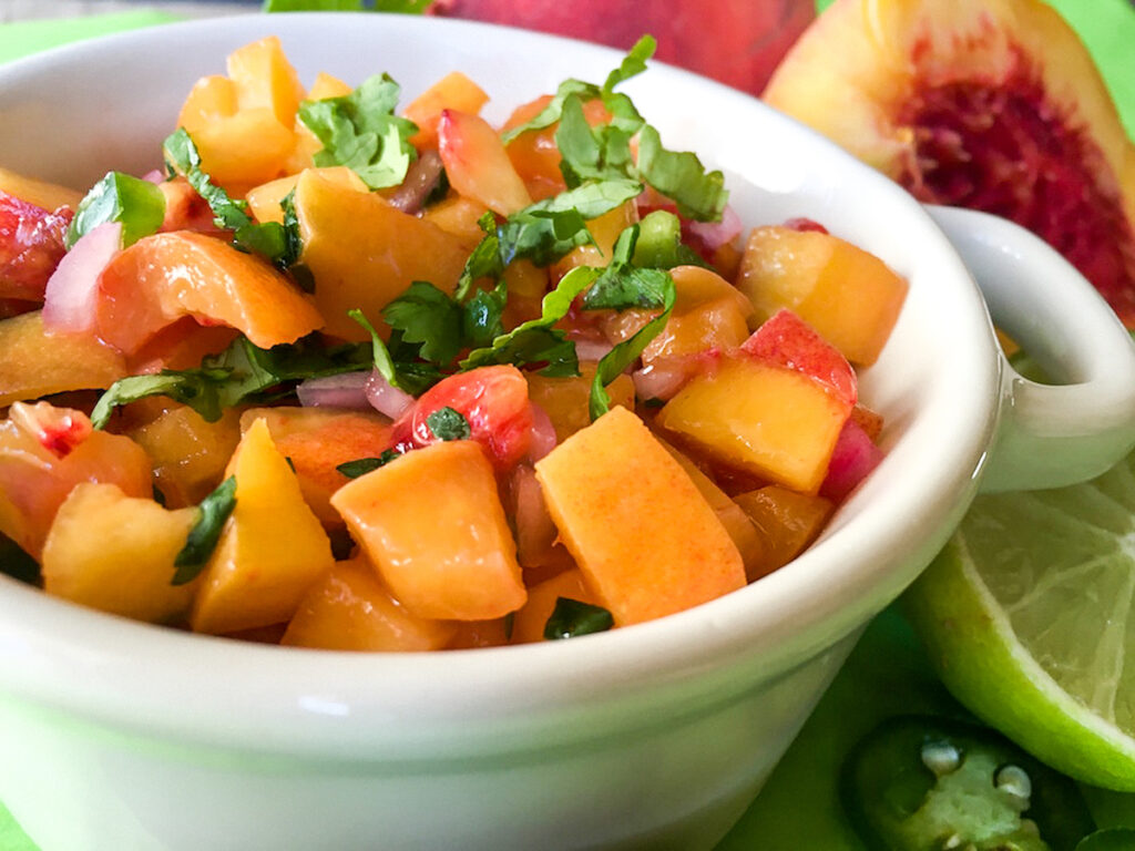 closeup of bowl of spicy peach salsa next to sliced peaches and limes