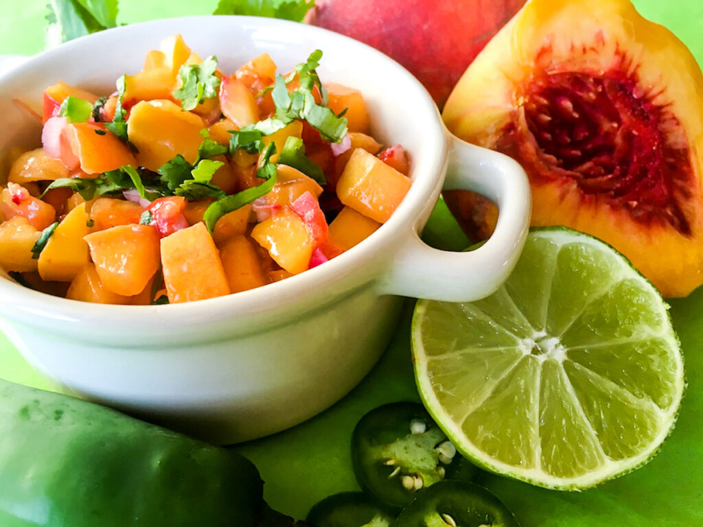 fresh peaches, sliced lime and jalapeno around a bowl of spicy peach salsa