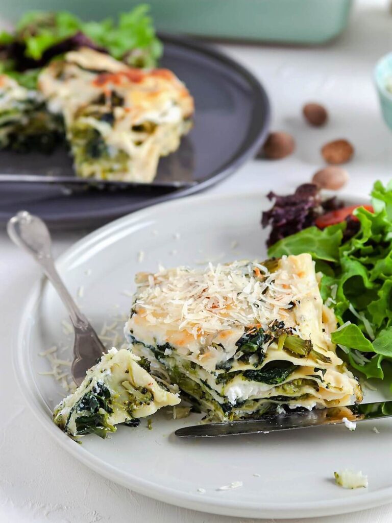 bite of spinach broccoli lasagna on a fork, next to a serving of spinach broccoli lasagna and green salad on a plate.