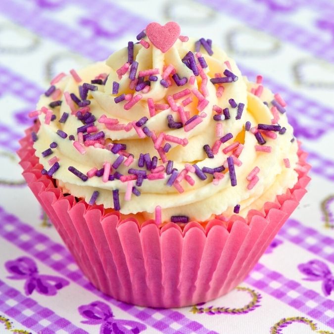 cupcake with buttercream frosting and pink, purple and yellow sprinkles in a pink cupcake liner on a festive purple and white patterned surface