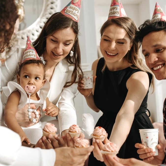 Three women and a baby with birthday party hats on all with cupcakes in their hands