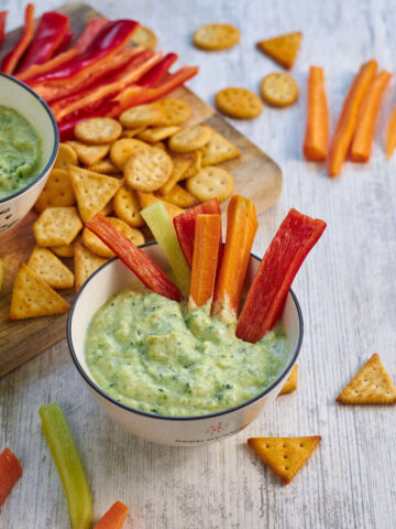 bowl of zucchini dip with sliced fresh bell peppers in it, in front of a cutting board with more fresh cut veggies and crackers