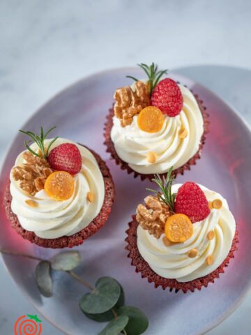 three yellow cupcakes with swirled buttercream frosting topped with raspberries, candied fruit and nuts