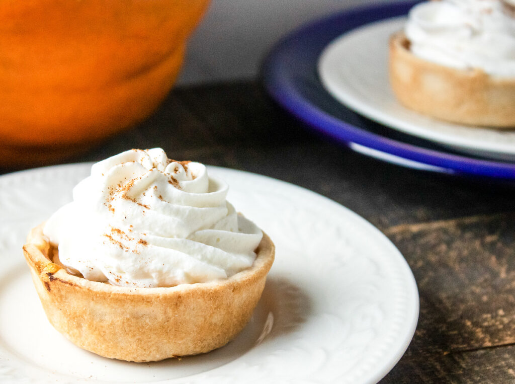 mini pumpkin pie from Flavor Portal recipe on a white plate in front of another plate with several more mini pumpkin pies on it