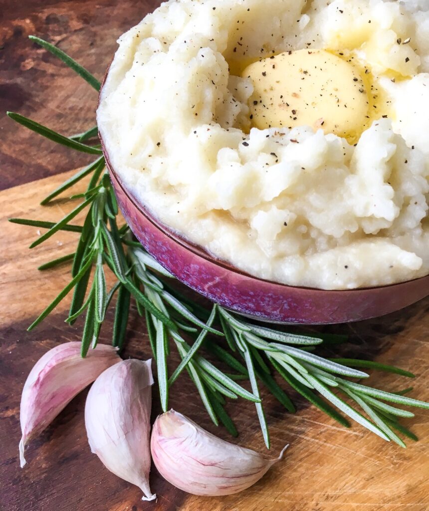 Instant Pot garlic mashed potatoes from Flavor Portal recipe in a bowl with melting butter pat next to garlic cloves and rosemary