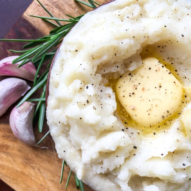 Instant Pot garlic mashed potatoes from Flavor Portal recipe in a bowl with melting butter pat next to garlic cloves and rosemary
