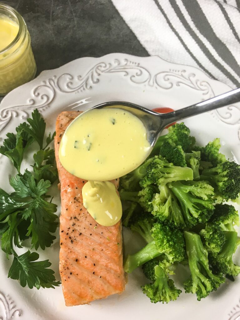 keto salmon with hollandaise sauce from Flavor Portal recipe on a white plate with steamed broccoli, cherry tomatoes and a parsley garnish with hollandaise sauce on the side