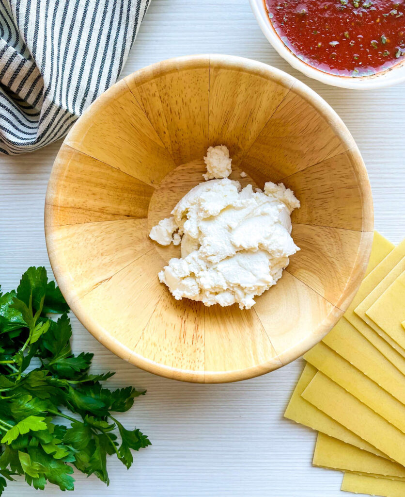 ricotta in a wooden bowl surrounded by parsley, pasta and tomato sauce for Spinach and Ricotta Lasagna Roll Ups from Flavor Portal recipe