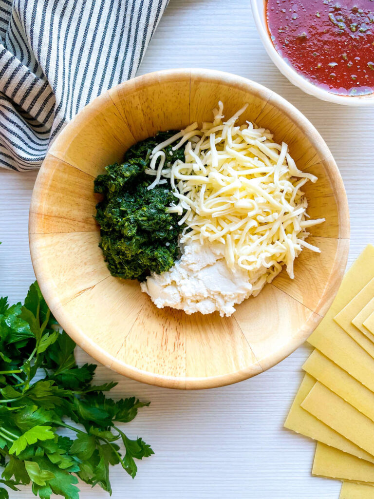 cheese added to spinach and ricotta in a wooden bowl surrounded by parsley, pasta and tomato sauce for Spinach and Ricotta Lasagna Roll Ups from Flavor Portal recipe