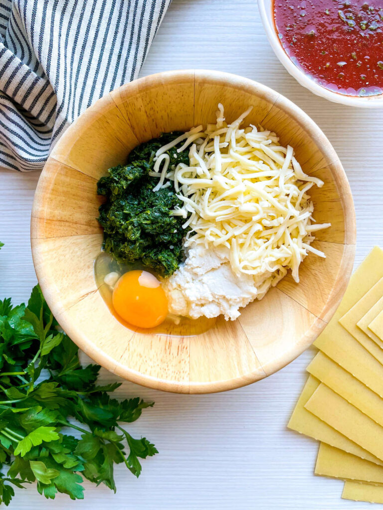 eggs added to spinach, cheese and ricotta in a wooden bowl surrounded by parsley, pasta and tomato sauce for Spinach and Ricotta Lasagna Roll Ups from Flavor Portal recipe