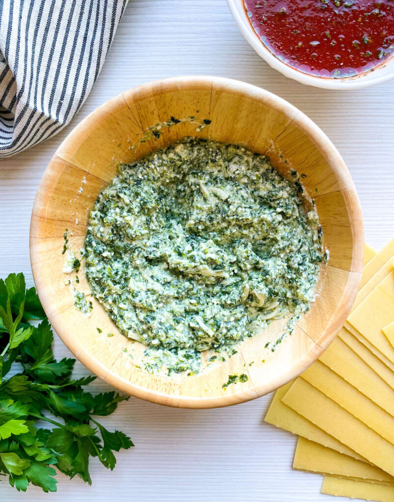 cheese and spinach mixture in a wooden bowl surrounded by parsley, pasta and tomato sauce for Spinach and Ricotta Lasagna Roll Ups from Flavor Portal recipe